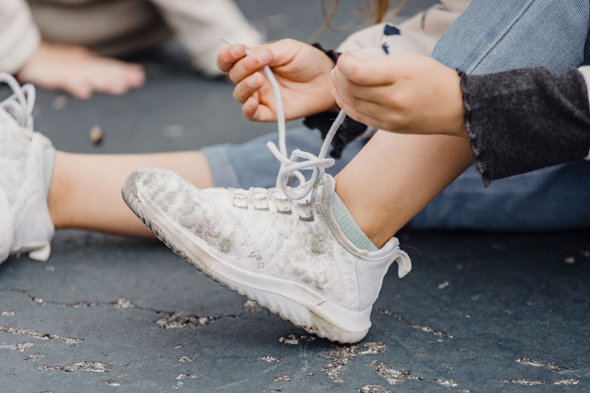 Un enfant assis par terre noue le lacet d’une basket blanche, vu en gros plan sur ses mains et sa chaussure.