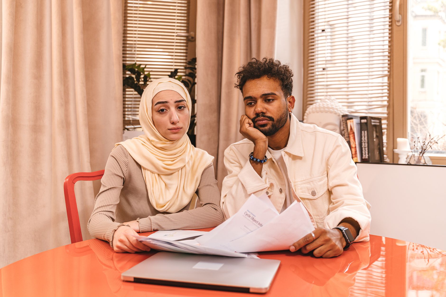Un homme et une femme assis à une table orange examinent des papiers, un ordinateur portable fermé devant eux, l’air préoccupé.