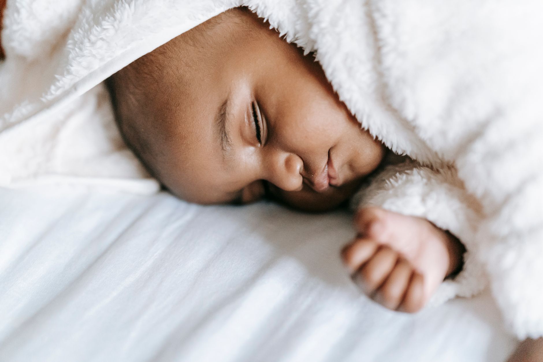 Un nourrisson dort sur un drap blanc, la tête et l’épaule recouvertes d’une couverture douce blanche, le petit poing fermé.